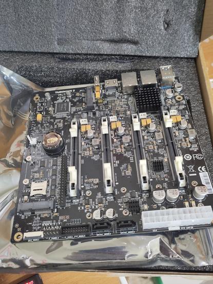 Square mainboard of a Turing Pi 2. Mainly black, with lots of scattered silver components. Stand outs are the slots for the add-on cards (running vertical) and the white power socket in the bottom right.
The board sits on the silvery anti-static bag, which sits on the black packaging foam.