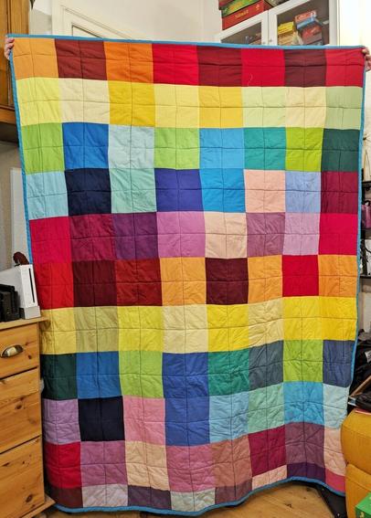 Living Room. A Person (you only see the fingers) holds a big quilt (145 x 190 cm). The quilt consists of big squares in uni colours. The colour change follows the rainbow scheme.