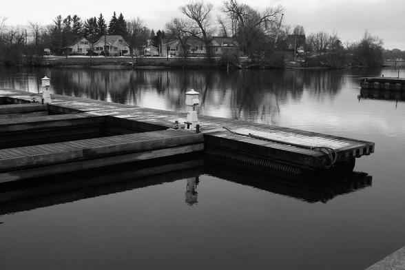 The calm water of a small lake shows the reflection of the floating docks and the trees on the distant shoreline. This is a black and white image.