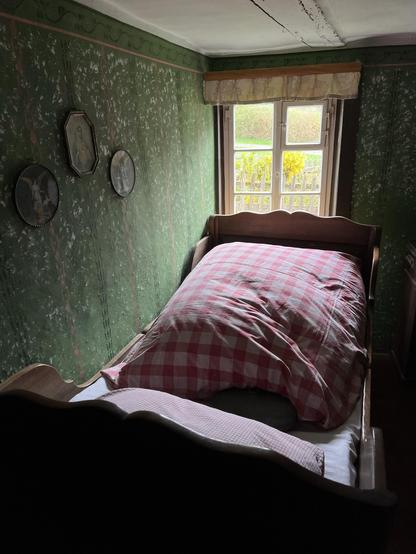In einem engen und niedrigen Raum steht ein altes Holzbett mit dickem Bettzeug vor einem kleinen Sptossenfenster. An der Wand zerschlissene, grüne Tapete. Über dem Bett hängen drei ovale Rahmen, mit Engeldarstellungen