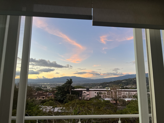 A window frames the pastel blues, oranges and yellows of sunset. Low buildings of the city are in the mid foreground. Beyond them rise the soft bluish gray of the mountains