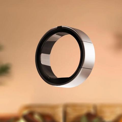 Photo of the Lotus Ring floating in space with a blurred background of a room with a couch. The Ring is circular and with a light brushed metallic outer surface and a black polished inner surface. Two slits are visible on the outer surface and a small black button is flush with the side ring. A small section of the inner surface is light gray.
