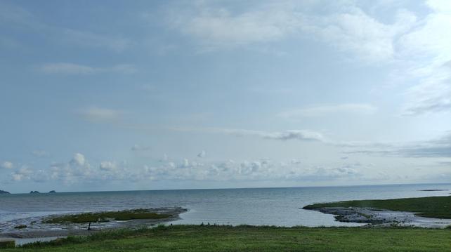 Vue paisible sur la mer depuis un rivage herbeux. Le ciel est légèrement nuageux, la lumière douce, et l’eau calme s’étend à perte de vue, bordée de petites îles à l’horizon