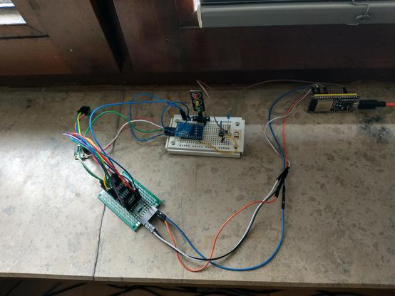 Photo of my little ESP32 tinker setup: an ESP32 NodeMCU development board rests on a marble window pane, along with a little white breadborad which holds a GPIO extender board and a custom made, green PCB which serves as I2C bus expander offering five I2C "ports". Two of these ports are being used by Temperature and Humidity sensors. All the different components are connected via jumper wires.