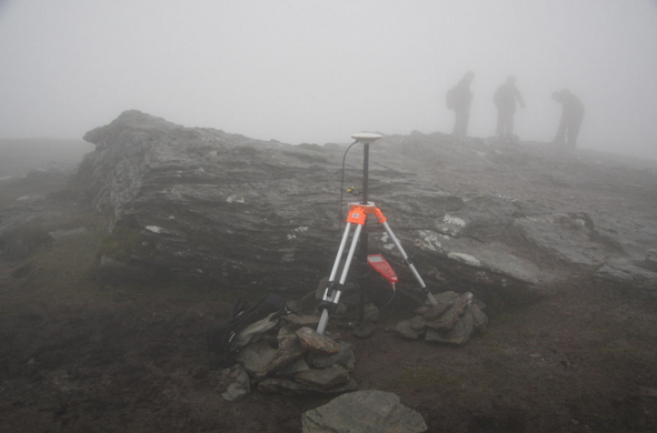 A photograph of the equipment set up on the summit for measuring the height of Ben Vane under very foggy conditions, with three figures barely visible through the fog.