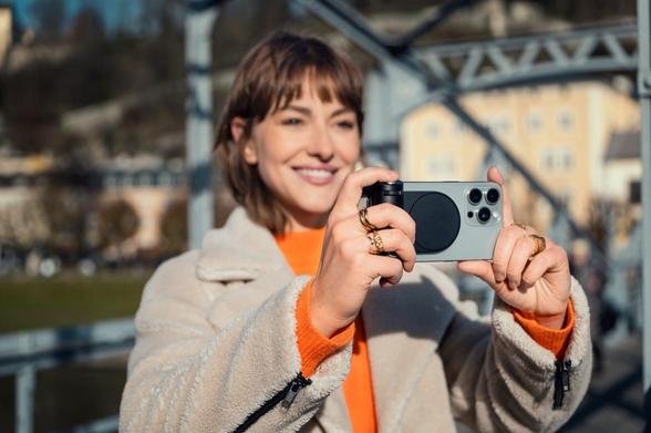 Eine lächelnde Person hält mit beiden Händen ein iPhone samt angebrachtem Leica Lux Grip fest und scheint etwas zu fotografieren. Im Hintergrund unscharf eine Stadtkulisse. Foto (c) Leica.
