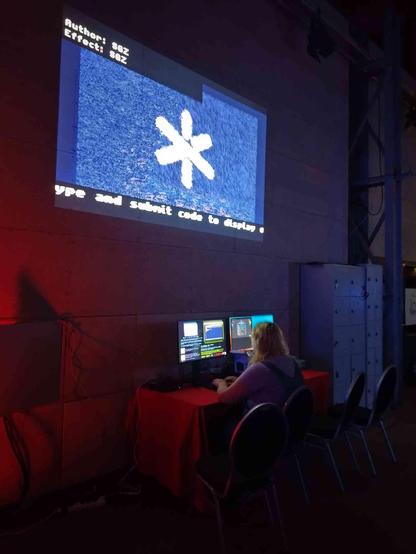 The ByteWall as an installation. A player is seated, writing code. Above, there's a projector running through a jukebox of the effects submitted so far.