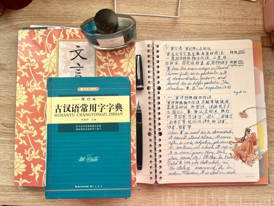 My Classical Chinese notebook with grammar explanations in modern Chinese followed by my English translation; stacked next to the textbook I’m following, a dictionary, my fountain pen, ink bottle, and a metal bookmark of a fantasy Chinese princess type maiden. The handwriting, dictionary and ink bottle are all teal green, while the textbook and the bookmark are coral pink.