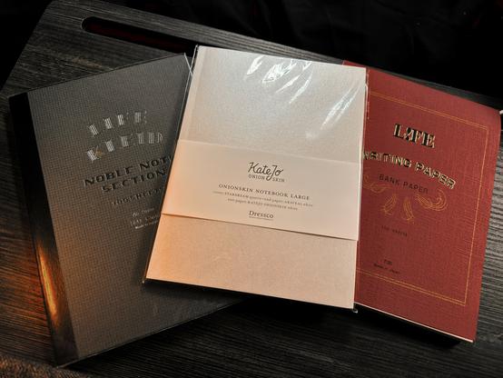 Top down photo of 2 A5 notebooks and a notepad: A Life Kleid Noble Note Section 2mm grid with a grey cover, a KateJo Onion Skin notebook with a creamy Stardream Quartz cover, and a Life Writing Paper - Bank Paper pad with a red cover. All sitting on a dark grey wooden tray.