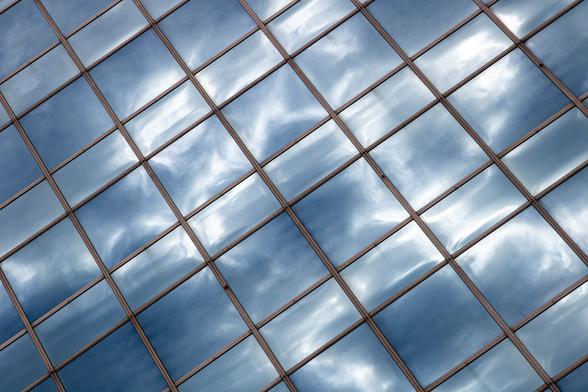 The facade of an office building, photographed diagonally from below. The building is made entirely of glass windows, rows of square windows alternating with rows of rectangular windows. The gray and white cloudy sky reflects in the windows. This reflection makes the windows appear convex, like a quilted blanket.