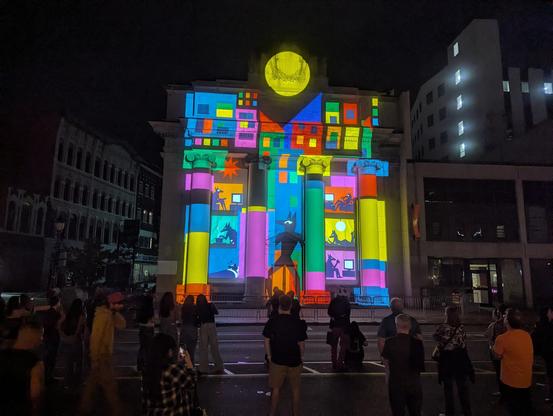 An animation projected onto a building, depicting the silhouettes of various bipedal wolf people as if in different apartments doing various things. One stands in the center looking around. The style features, bright colors, and very blocky artwork.