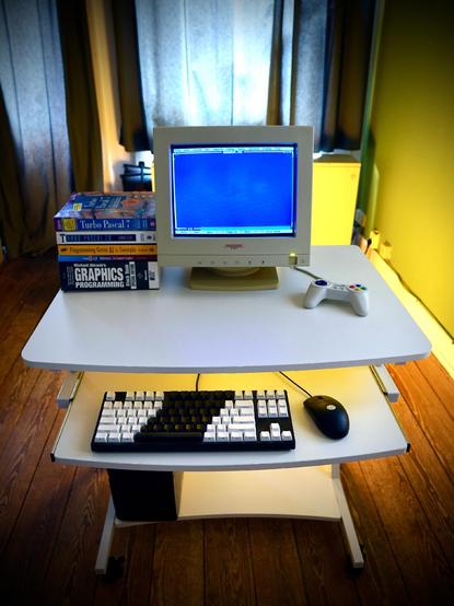 Small computer desk with a 14" crt monitor on top, a black-and-white mechanical keyboard and a black mouse on the keyboard tray, and a black mini tower on the bottom shelf. Next to the monitor is a stack of books (Graphics Progamming Black Book, Mathmatics for Computer Graphics, Programming Game AI by Example, and two Turbo Pascal 7 books)