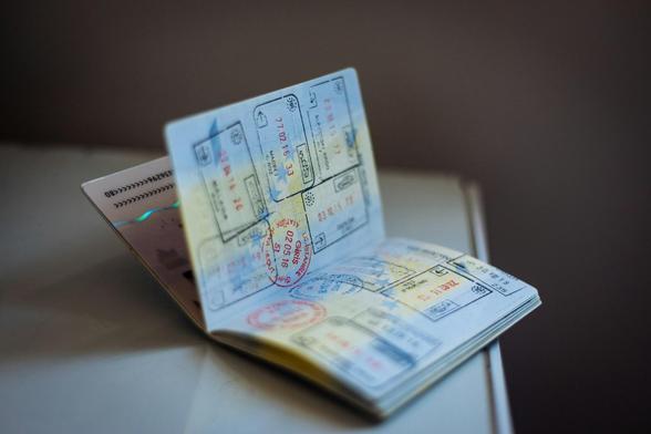 An open passport displaying multiple colorful visa stamps and entry/exit stamps on its pages. The passport appears to be resting on a white surface, with various stamps visible including what looks like arrival and departure dates. The stamps show different colors including red, blue, and black ink, indicating travel to multiple countries. The passport's pages are fanned open to show the collection of travel documentation.