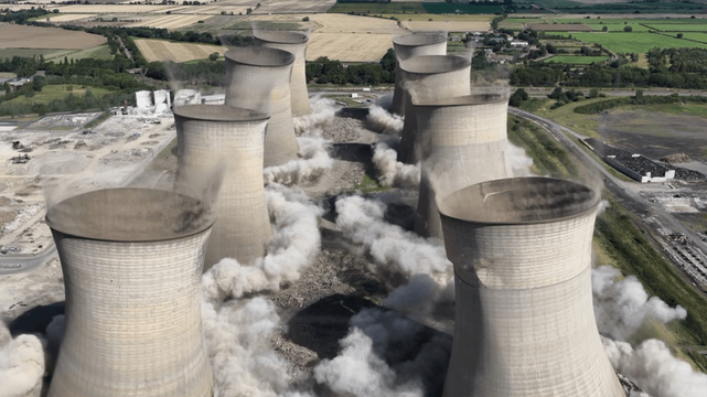 Aerial view looking down on two rows containing four cooling towers each, mid-demolition. Smoke and dust billow out from the base of each tower.