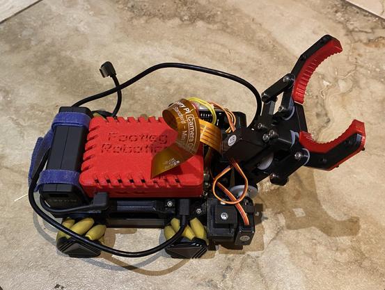 A small mechanum wheeled robot with a barrel grabbing claw on the front. A Raspberry Pi 5 is mounted in a custom 3D printed case.