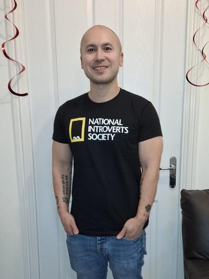 A photo of me stood in front of a door wearing a black T-shirt that has the words "National Introverts Society" printed on the front