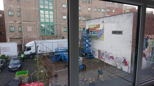 Looking out on an industrial back street in central Liverpool. There's a surface level car park next to a flat-roofed brick warehouse.

The car park is devoid of cars and instead populated with a couple of blue cherry pickers and is surrounded by a high "fence" made of scaffold poles.

The end wall of the warehouse is painted white with graffiti along the bottom, and there are two painters on a scissor lift finishing off an old-style advert imploring us to "Ask for Mendos soap".

In the background are two white trucks with Panalux logos on the side.
