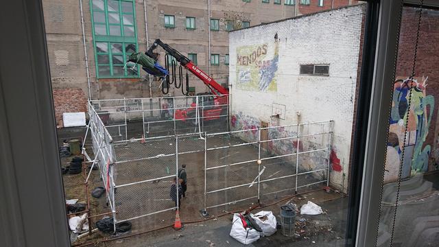 A half-basketball court with chain-link fencing round three sides; the fourth side is the wall of a brick warehouse.

There's a faded sign for Mendos soap painted on the side of the building, and graffiti along the bottom of the wall; and bits of stained paintwork dotted here and there.

All of that has been carefully painted by the set artists, and the whole fence has been constructed and painted over the last day or two.

There's a firefighting hose running along the edge of the court, which is actually for creating the clouds of steam to make the area look like New York (well, Gotham City) and in the background is a large red teleloader which has a seriously large lighting rig on it.