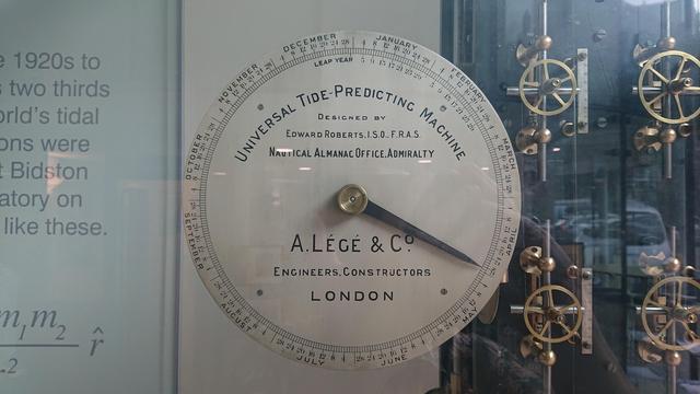 A dial showing the months and days of the year around its edge and a pointer indicating April 28th.

The centre is labelled "Universal Tide-Predicting Machine.  Designed by Edward robers, I.S.O., F.R.A.S.  Nautical Almanac Office, Admiralty.  A. Légé & Co.  Engineers, Constructors.  London"

A few brass wheels and mechanisms can be seen to the right.