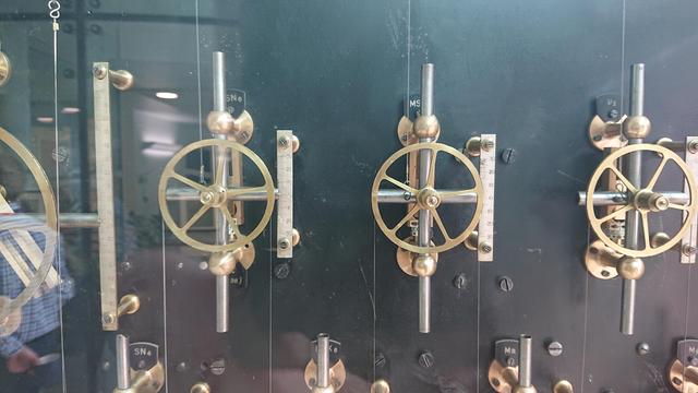 A closer shot of some of the brass pulleys and mechanisms of the machine.  There's a wire running over each one, that wends its way around the whole machine, and they're all labelled with their particular "variable" that they add to the result: SN4, V2, Mm, etc.

The linkage that each pulley is mounted on has a horizontal steel bar with a pointer that indicates its value on a scale mounted to the right of the mechanism; and there's a vertical steel bar to let the pulley-and-horizontal-bar slide up and down.  Behind all that are brass screw adjustments to adjust the linkages to set the coefficient of that variable.