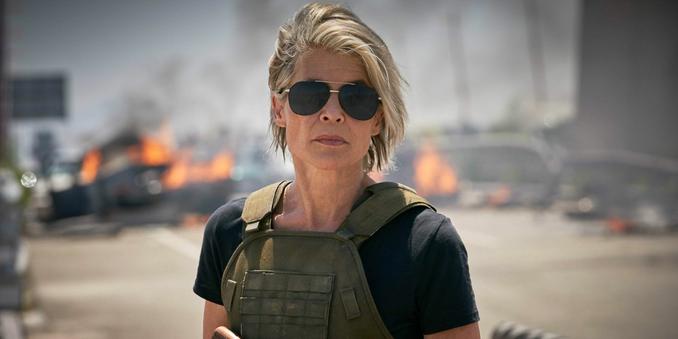 An older woman (Sarah Connor of the Terminator movies) standing on a highway with wrecked and flaming cars. She has a bulletproof green vest and is wearing dark aviator sunglasses, and has a grim expression.