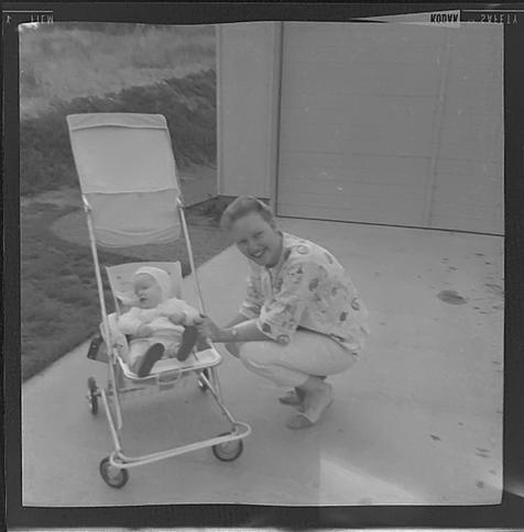 Black and white image of a woman squatting down by a baby stroller in a suburban driveway. The infant is ME!