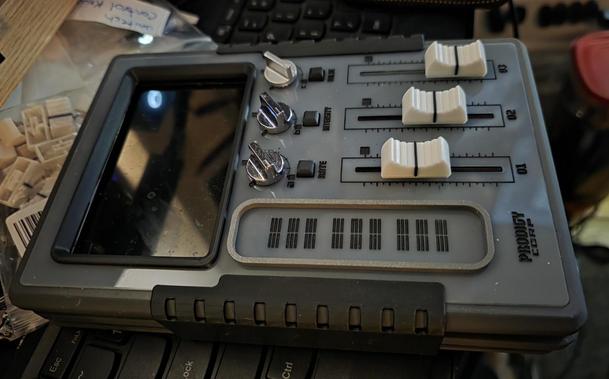 A replica of the hybrid control device from Alien Earth. It is a rectangular gray device with three faders, three buttons, three knobs, and a screen at the top