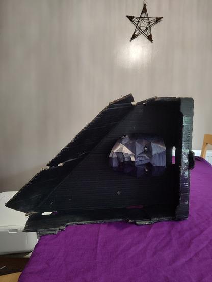 A black painted cardboard box with the top left corner cut diagonally at 45 degrees sits on a purple table cloth in front of a beige wall with a pen tangle ornament made of of twigs hanging on it in the background. Mounted inside the box is a stern looking black angular 3d printed face. It's on its side, chin to the left, because the box won't stand up the right way on a table because of the cut corner.