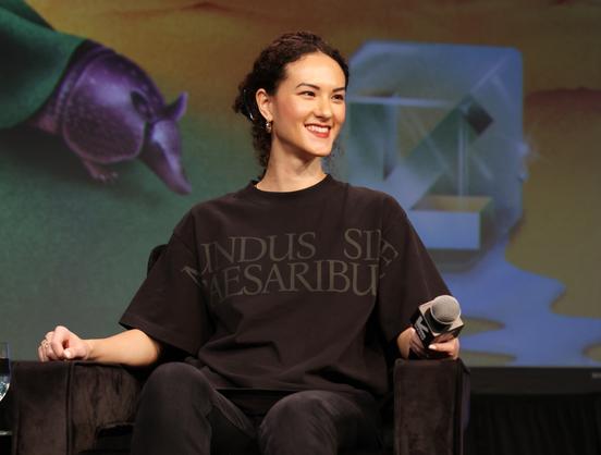 Woman in black t-shirt reading 'mundus sine caesaribus' sits in chair onstage.