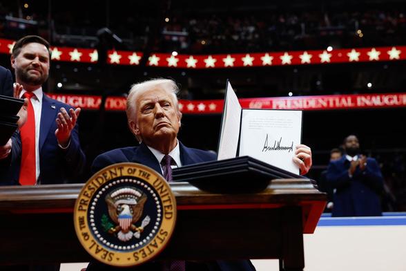 U.S. President Donald Trump sitting at a desk with the presidential seal, holds up an executive order after signing it, with Vice President JD Vance standing behind him, during an indoor inauguration parade at Capital One Arena on January 20, 2025 in Washington, DC.