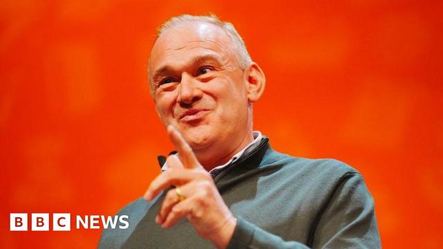 Ed Davey smirks and raises a finger to make a point against a bright orange background, on stage at the Lib Dem conference
