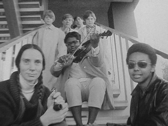 A 16mm black-and-white still of film from the performance of Robert Moran’s “Jewel Encrusted Butterfly Wing Explosions” by the Oakland Symphony’s Youth Chamber Orchestra showing members of the Youth Chamber Orchestra playing their instruments in various public places in San Francisco and Oakland, from the beach, to the Legion of Honor, in trees, in the middle of streets and on staircases, as well as in front of the Oakland Public Museum.