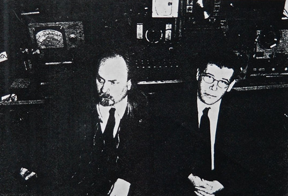 A black-and-white copier photo of the duo The Hearing Trumpet wearing their trademark dark suits with white shirts and black ties while sitting in front of their recording gear.