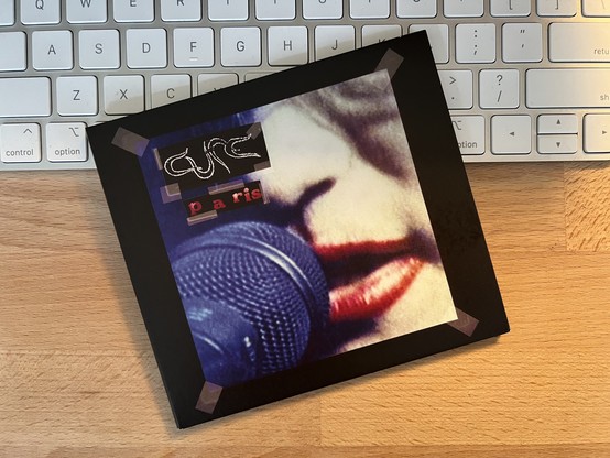 A photo of the album cover for “Paris” by The Cure features a closeup image of Robert Smith’s face with his bright red lipstick in front of a microphone. The colors are kinda washed out giving it a dreamy feeling.
