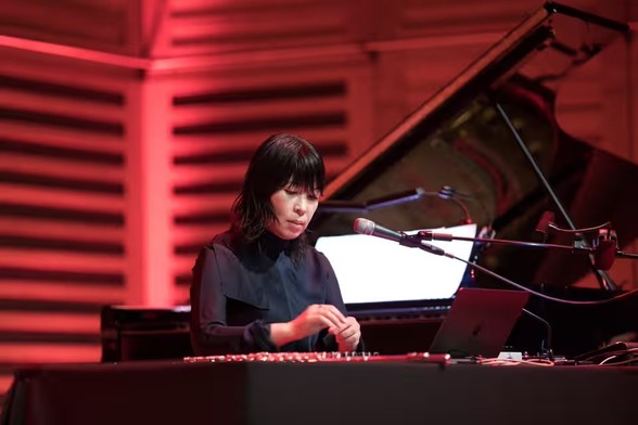 A photo of Eiko Ishibashi performing at the London jazz festival in 2022. She is sitting at a table, bathed in red light, with what looks like an electric guitar and effects pedals. Behind her is a grand piano. Photo by Teri Pengilley/The Guardian.