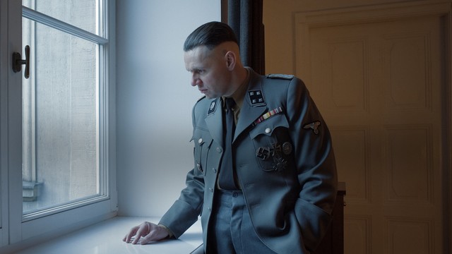 A movie still depicting Rudolf Höss, commandant of Auschwitz, standing in his Nazi uniform while looking out a window from his office. He spends a lot of time pondering the efficiency of his “work” to carry out Hitler’s final solution.
