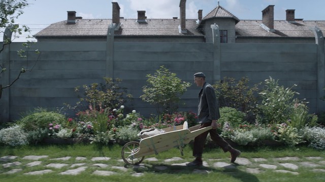 A movie still depicting a prisoner of Auschwitz pushing a wheelbarrow with sacks through the back yard of the commandant’s villa adjoining the camp’s barracks.
