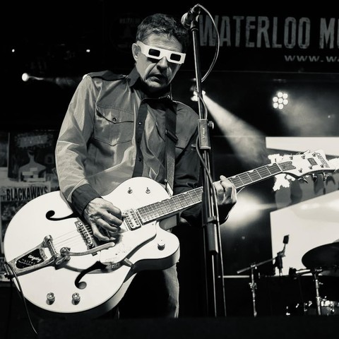 A black-and-white photo of Graeme Naysmith playing what looks like a classic Gretsch hollow body guitar. He is wearing paper 3-D vision glasses.