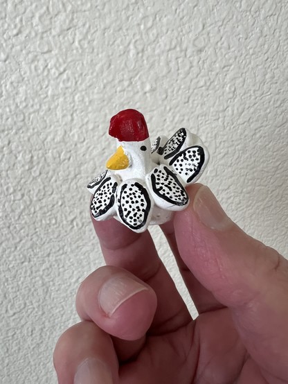 A photo of a hand holding a very small chicken figurine with a friendly demeanor. It’s painted white with black specks on its feathers, a yellow beak, and a red crown.