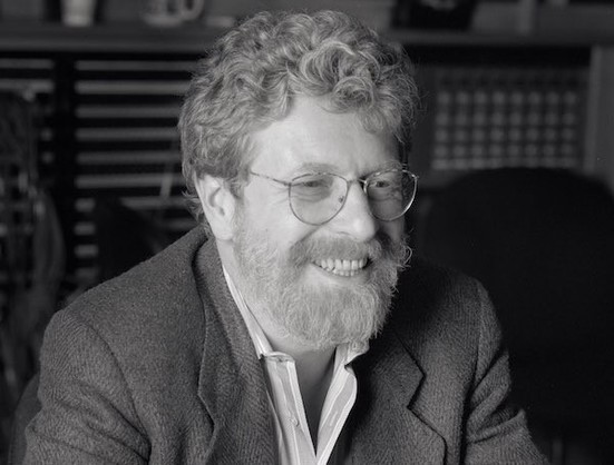 A black-and-white photo of the Blue Note historian and producer Michael Cuscuna, circa mid-1990s. He is smiling, has wire-rimmed glasses with a beard, and is wearing a tweed suit coat with his shirt collar unbuttoned.