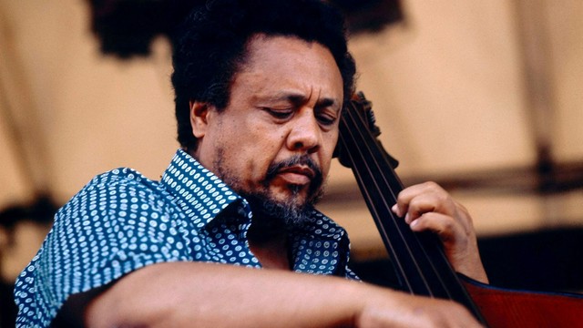 A color photo, circa 1960s, of Charles Mingus wearing a blue polka dot button down shortsleeved shirt and playing bass.