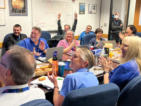 A color photo of members of the Voyager flight team celebrate with donuts in a conference room at NASA’s Jet Propulsion Laboratory on April 20.