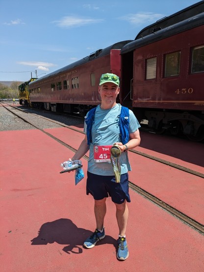 Runner standing in front of a historic train wearing a race bib and hydration vest, holding a race medal and T-shirt. Sunny day 
