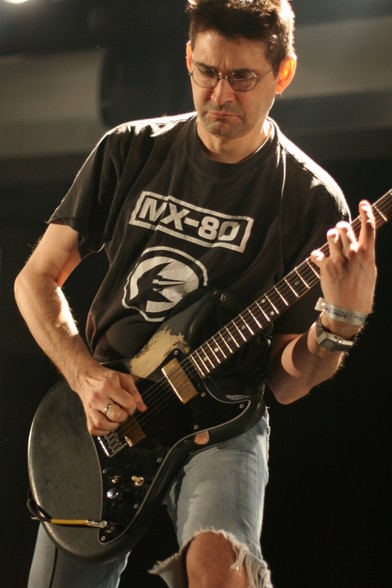 A color photo of Steve Albini, wearing a black MX-80 t-shirt while playing guitar during a live Shellac concert.