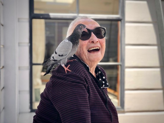 A color photo of Barbara Stauffacher Solomon in 2023. She is smiling, standing outside her home in San Francisco, wearing an eggplant purple blouse with black stripes and sunglasses. Oh yeah, and there’s a pigeon perched on her shoulder! 📸 by her daughter, Nellie King Solomon.