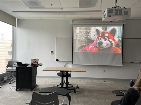 A photograph of a classroom. A slide from a presentation titled “The Furry Fandom and Conceptual Metaphors in Identity Craft” is being projected on the screen. The background of the slide is a pair of red panda fursuiters.