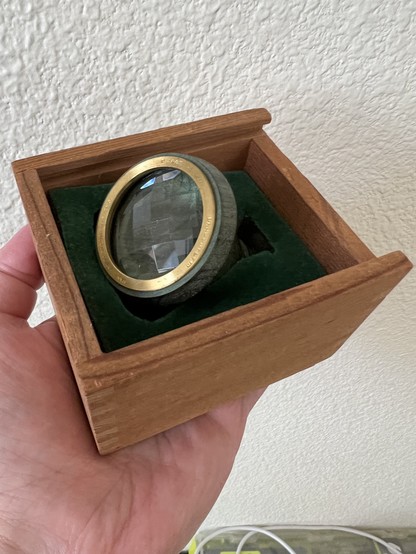 A closeup color photo of me holding my green birch wood Dragonfly kaleidoscope in the palm of my hand wherein you can see it snuggled in the plain birch wood box (lined with green felt) I keep it in.