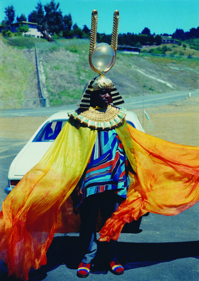 A photo of Sun Ra wearing one of his colorful outfits — striped in blues and reds, with a long, flowing satin cape in yellow to red, and his signature crown of the Egyptian sun god symbol — taken while filming his docudrama,  “Space Is the Place” (1974). 📸 courtesy of the collection of John Corbett and Teri Kapsalis.