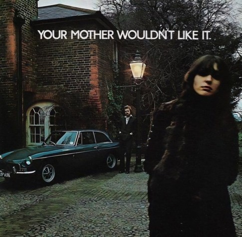 Like an album cover, but it’s an advert for an MGB in 1972. It’s an overcast day. There’s a woman wearing a black leather overcoat in the foreground. Parked in front of a brick mansion is a blue sports car with a man wearing a black suit and white button down shirt standing next to it. The caption reads: “Your Mother Wouldn’t Like It”