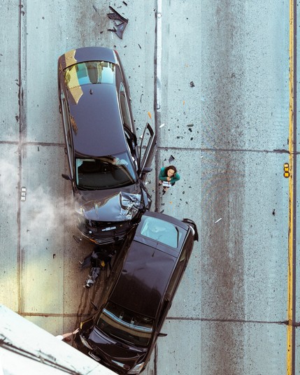 Imagine you are peering straight down from atop an overpass at what looks like the immediate aftermath of three car pileup. Two black cars with crumpled ends are visible. There’s smoke rising from a radiator, scattered debris on the road, and a woman wearing a green coat looking up at the camera.